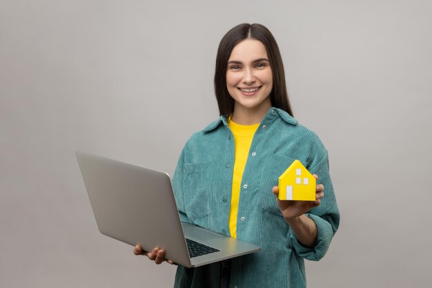 Immobilienmaklerin steht mit Papierhaus und Laptop in der Hand und hilft bei der Miete