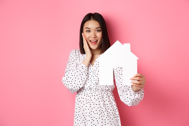 Imobiliária. Mulher asiática adulta procurando por casa, segurando o modelo da casa e sorrindo, promoção da corretora, em pé sobre a rosa
