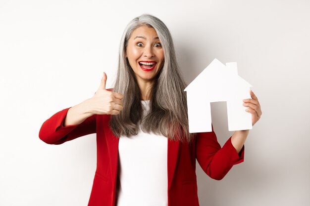 Imobiliária. Retrato do corretor feminino asiático mostrando o polegar para cima e o recorte da casa de papel, recomendando a agência para comprar a propriedade, em pé feliz sobre o fundo branco.