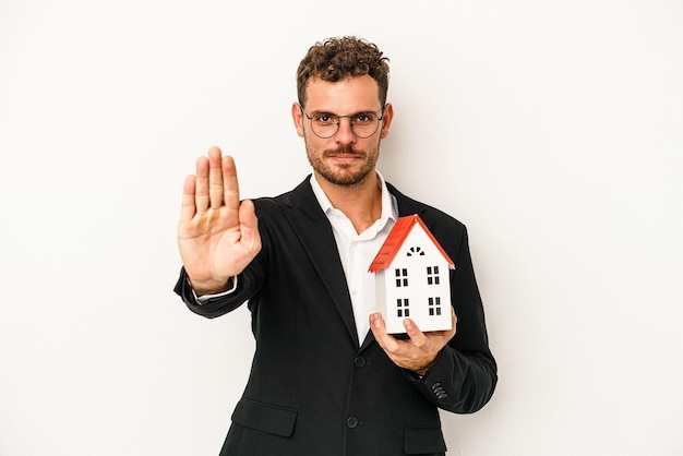 Imobiliário caucasiano jovem segurando uma casa de brinquedo isolada no fundo branco em pé com a mão estendida, mostrando o sinal de pare, impedindo você.