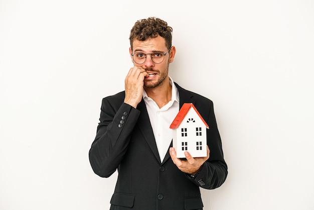 Imobiliário caucasiano jovem segurando uma casa de brinquedo isolada no fundo branco, roer as unhas, nervoso e muito ansioso.