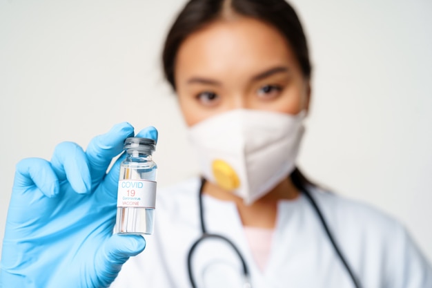 Impf- und Covid-19-Präventionskonzept. Nahaufnahme eines asiatischen Arztes in Atemschutzmaske zeigt eine Flasche mit Coronavirus-Impfstoff, die auf weißem Hintergrund steht