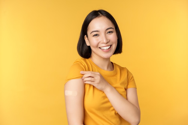 Impf- und covid19-Pandemiekonzept Fröhliches und gesundes asiatisches Mädchen, das nach der Impfung vor gelbem Hintergrund des Coronavirus mit Bandhilfe auf ihre Schulter zeigt