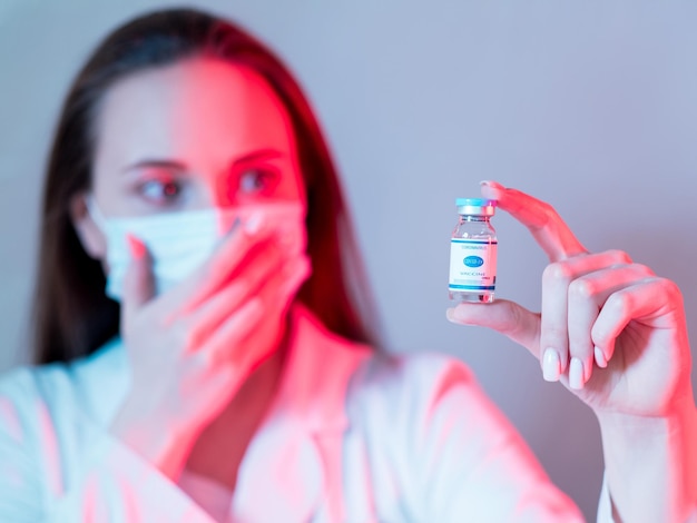 Impfgefahr Covid19-Immunisierung Innovative Medizin Schockierte Ärztin in Gesichtsmaske testet die Fläschchendosis in rotem Neonlicht isoliert auf unscharfem violettem Hintergrund