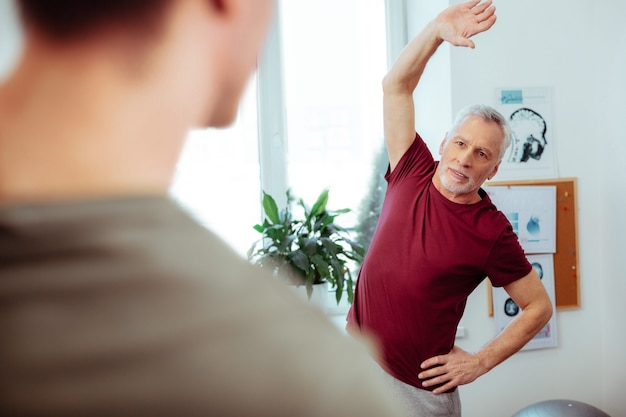 Importancia del deporte. Anciano agradable haciendo ejercicios físicos mientras tiene un entrenamiento deportivo