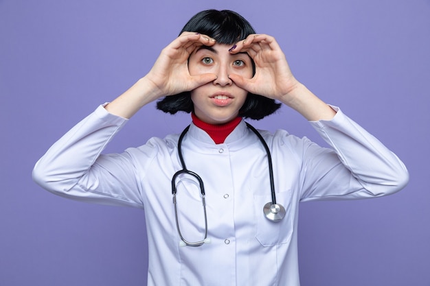 Impresionada joven bastante caucásica en uniforme médico con estetoscopio a través de los dedos