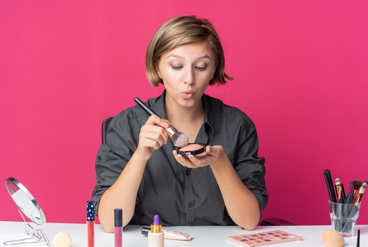 Foto impresionada joven bella mujer se sienta a la mesa con herramientas de maquillaje con rubor en polvo con pincel