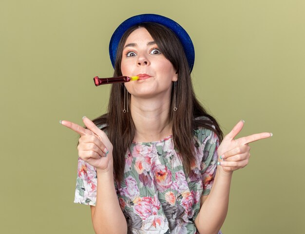 Impresionada joven hermosa mujer vistiendo gorro de fiesta soplando silbatos de fiesta en los lados aislados en la pared verde oliva