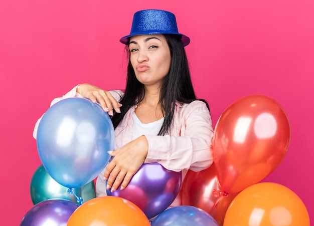 Impresionada joven hermosa con sombrero de fiesta de pie detrás de globos