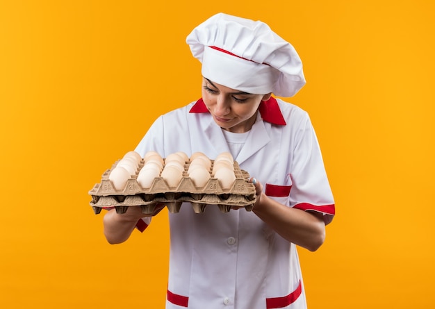 Impresionada joven hermosa en uniforme de chef sosteniendo y mirando lote de huevos