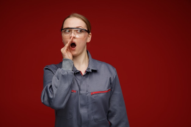 Impresionada joven ingeniera rubia vestida con uniforme y gafas de seguridad manteniendo la mano cerca de la boca mirando al lado susurrando