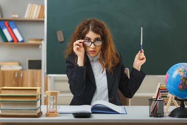 impresionada joven maestra con anteojos apuntando a la pizarra con un puntero sentado en el escritorio con herramientas escolares en el aula