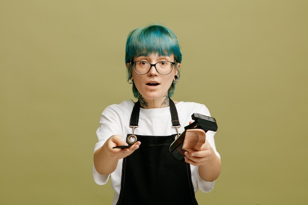 Impresionada joven peluquera con gafas uniformes mirando a la cámara estirando peines y spray para el cabello hacia la cámara aislada en un fondo verde oliva