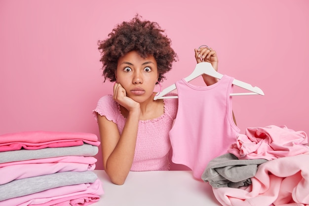 Impresionada mujer afroamericana ordena ropa sostiene ropa en percha selecciona ropa del armario para donación en tienda benéfica rodeada de lavandería aislada en rosa