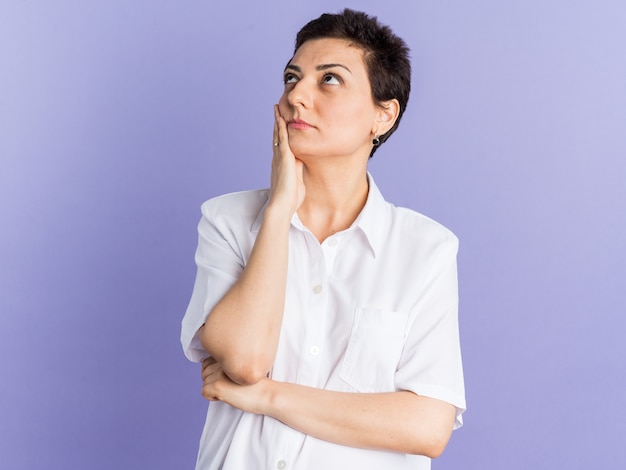 Impresionada mujer de mediana edad manteniendo la mano en la cara mirando hacia arriba