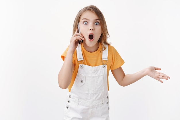 Impresionada niña rubia carismática, niño hablando por teléfono, llamando a un amigo chismeando, mirando sorprendido y asombrado con la boca abierta, gesticulando con la mano levantada cuestionada, usando el teléfono inteligente y contando rumores jugosos