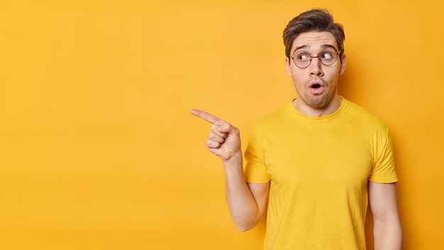 Impresionado, un hombre adulto atónito señala con el dedo hacia afuera y demuestra un anuncio impresionante que contiene el aliento, no puede creer que sus ojos usen anteojos redondos transparentes y una camiseta casual aislada sobre una pared amarilla