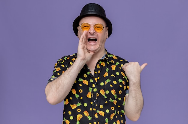 Impresionado hombre adulto con sombrero de copa negro con gafas de sol manteniendo la mano cerca de la boca y apuntando al lado