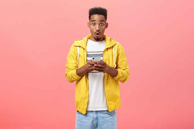 Impresionado hombre afroamericano entusiasta con labios plegables de corte de pelo rizado en sonido wow sosteniendo un teléfono inteligente emocionado y emocionado con características geniales del dispositivo o una nueva aplicación sobre una pared rosa