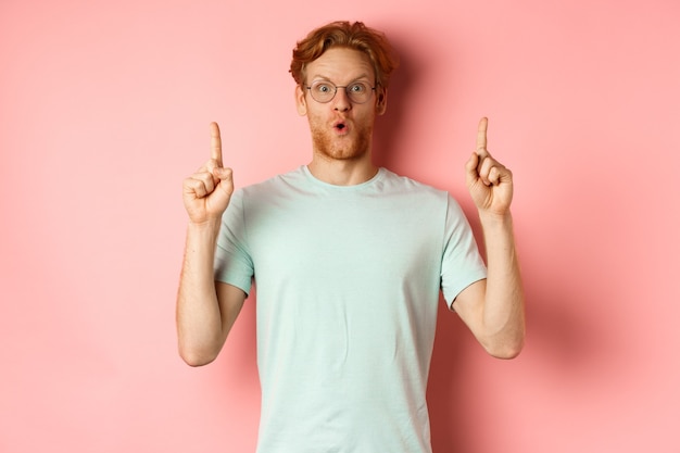 Impresionado hombre caucásico con pelo pelirrojo con gafas y camiseta diciendo wow y señalando con el dedo ...