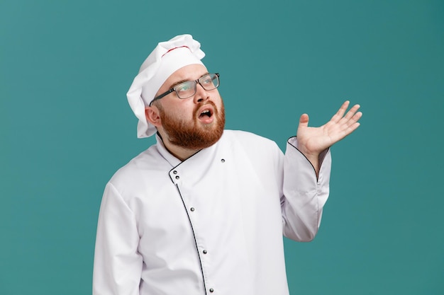 Impresionado joven chef masculino con anteojos uniforme y gorra mostrando la mano vacía mirando al lado aislado en el fondo azul