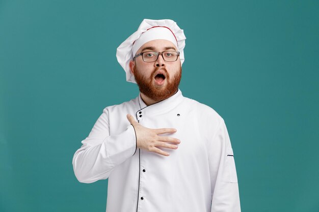 Impresionado joven chef masculino con uniforme de anteojos y gorra mirando a la cámara manteniendo la mano en el pecho aislado en el fondo azul