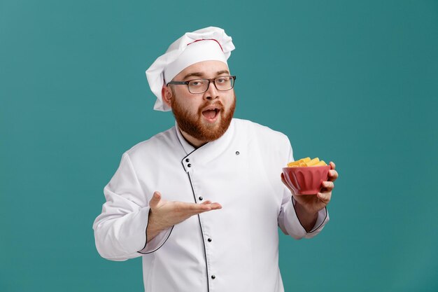 Foto impresionado joven chef masculino con uniforme de anteojos y gorra sosteniendo un tazón de macarrones apuntando con la mano mirando a la cámara aislada en el fondo azul