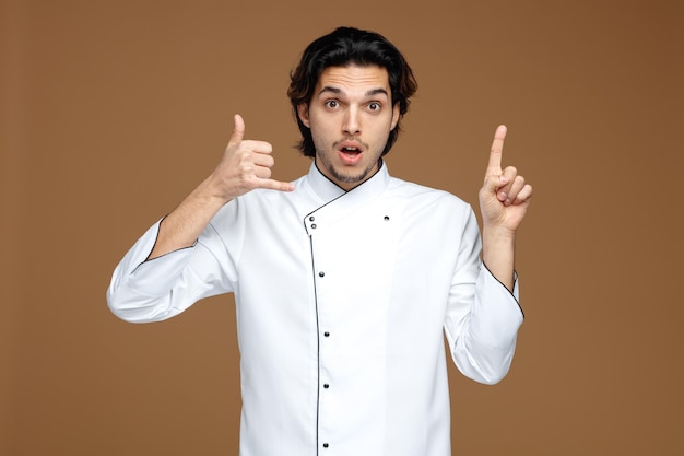 Impresionado joven chef masculino vistiendo uniforme mostrando gesto de llamada mirando a la cámara apuntando hacia arriba aislado sobre fondo marrón