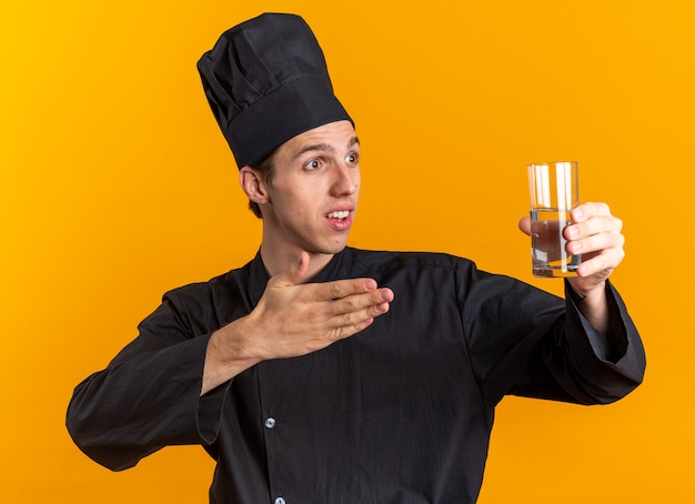 Impresionado joven cocinero rubio en uniforme de chef y gorra mirando a un lado estirando y apuntando al vaso de agua aislado en la pared naranja