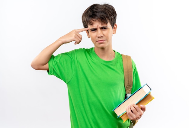 Impresionado joven escolar con mochila sosteniendo libros poniendo el dedo en el templo aislado en la pared blanca