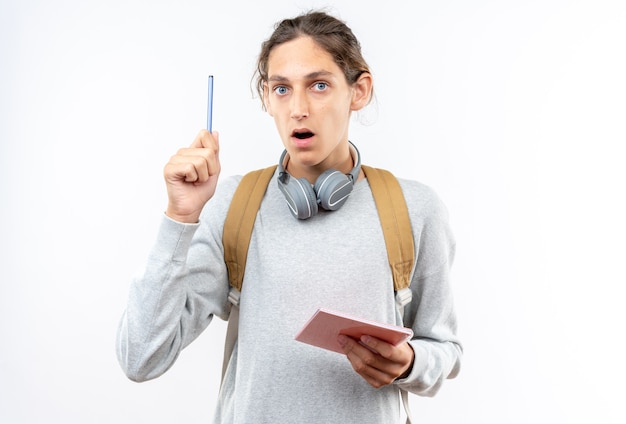 Impresionado joven estudiante con mochila con auriculares en el cuello sosteniendo el cuaderno levantando la pluma aislada en la pared blanca