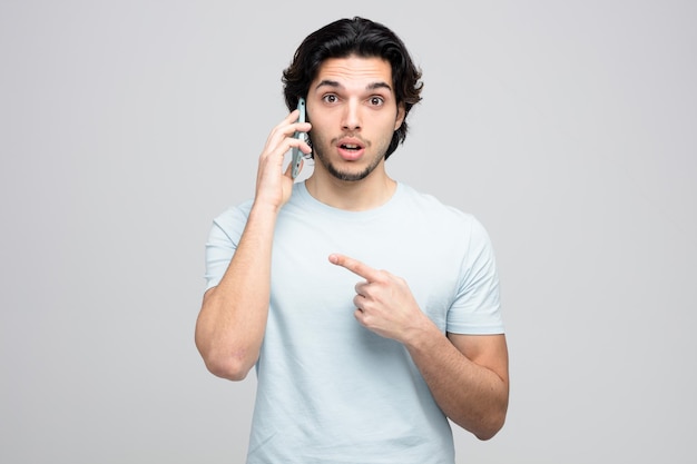 Impresionado joven guapo mirando a la cámara mientras habla por teléfono apuntando al lado aislado sobre fondo blanco.