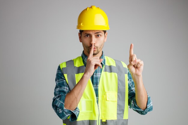 Impresionado joven ingeniero masculino con casco de seguridad y uniforme mirando a la cámara mostrando un gesto de silencio apuntando hacia arriba aislado en fondo blanco