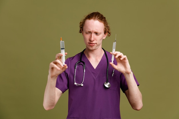 Impresionado joven médico masculino vistiendo uniforme con estetoscopio sosteniendo jeringa aislado sobre fondo verde