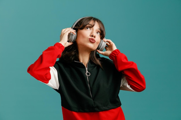 Impresionado joven mujer caucásica usando y agarrando auriculares mirando hacia arriba aislado sobre fondo azul.