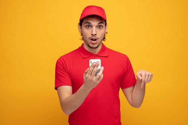 Impresionado joven repartidor con uniforme y gorra sosteniendo un teléfono móvil mirando a la cámara apuntando hacia abajo aislado en un fondo amarillo