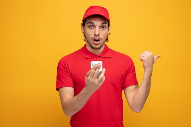 Impresionado joven repartidor con uniforme y gorra sosteniendo un teléfono móvil mirando a la cámara apuntando hacia un lado aislado de fondo amarillo