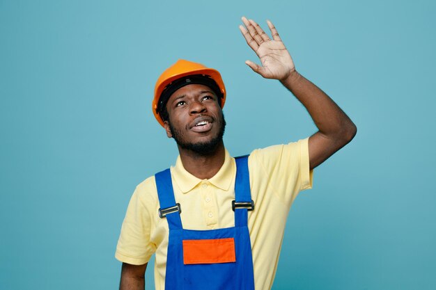 Impresionado mano levantada joven constructor afroamericano en uniforme aislado sobre fondo azul.