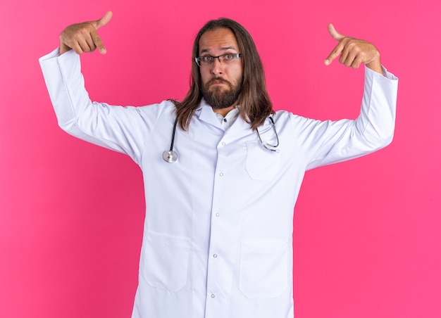 Impresionado médico varón adulto con bata médica y un estetoscopio con gafas mirando a la cámara apuntando al espacio frente a él aislado en la pared rosa