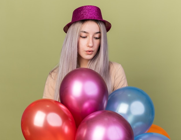 Impresionado mirando hacia abajo hermosa joven vistiendo gorro de fiesta de pie detrás de globos