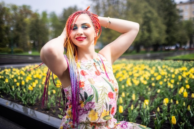 Impresionante chica con trenzas de arcoíris y expresivo maquillaje con purpurina. Se sienta y disfruta del cálido clima primaveral en un parque verde con grandes camas de tulipanes.