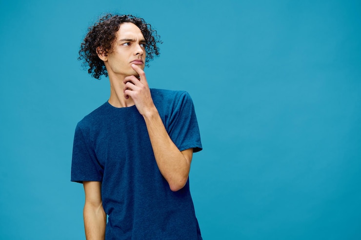 Foto impresionante chico caucásico bronceado rizado fresco en camiseta básica mostrar lugar libre posando aislado sobre fondo azul concepto de estilo de vida y emociones buena oferta con espacio de copia para anuncio