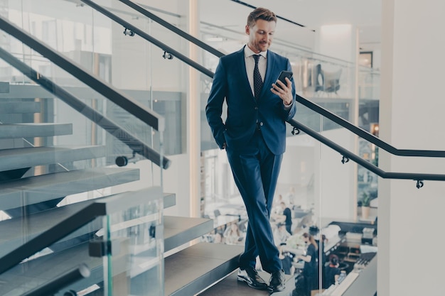 Impresionante empresario en traje elegante formal de pie en la escalera en el vestíbulo de la oficina con teléfono inteligente