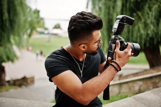 Impresionante y hermoso fotógrafo de barba alta con gafas y camiseta negra con cámara profesional en las manos