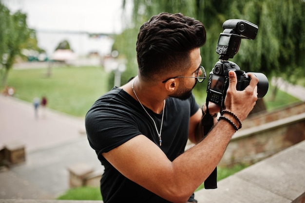 Impresionante y hermoso fotógrafo de hombre macho de barba árabe alto con gafas y camiseta negra con cámara profesional en las manos