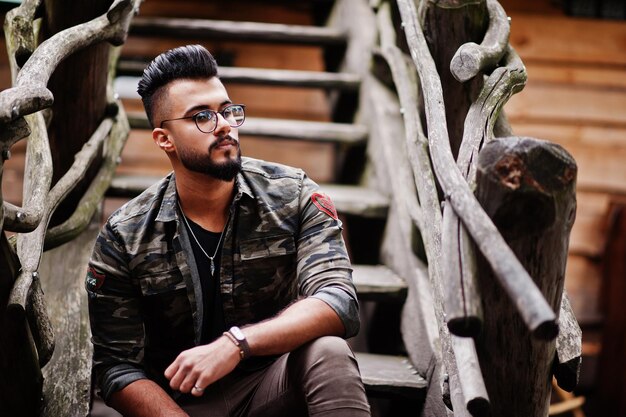 Impresionante hermoso hombre macho alto de barba árabe con gafas y chaqueta militar posado al aire libre contra escaleras de madera