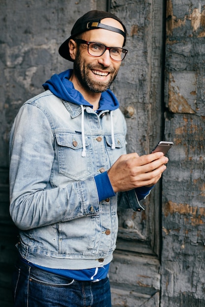 Impresionante hombre con barba de ojos azules y amplia sonrisa con camisa de mezclilla y anteojos sosteniendo un teléfono inteligente aislado sobre el fondo de la pared vieja Hombre complacido con ropa elegante usando un teléfono celular