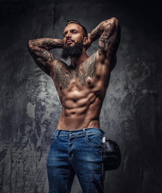 Impresionante hombre barbudo tatuado con el torso desnudo posando sobre fondo gris.