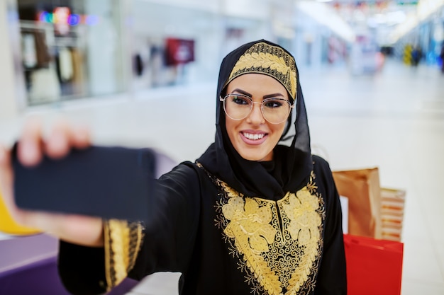 Impresionante joven árabe en ropa tradicional con una hermosa sonrisa con dientes de pie en el centro comercial y tomando selfie para las redes sociales. Generación millennial.