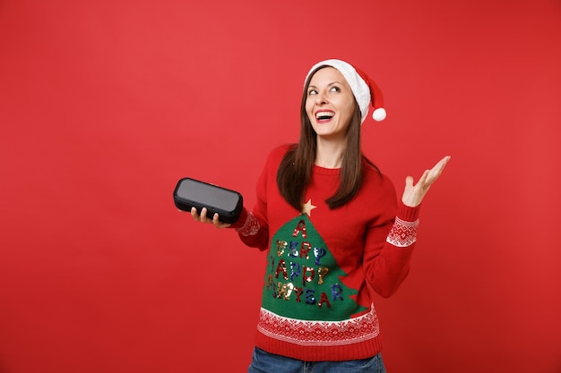 Impresionante joven Santa mirando hacia arriba, sosteniendo el altavoz de música bluetooth inalámbrico portátil aislado sobre fondo rojo brillante. Feliz año nuevo 2019 celebración concepto de fiesta navideña. Simulacros de espacio de copia.